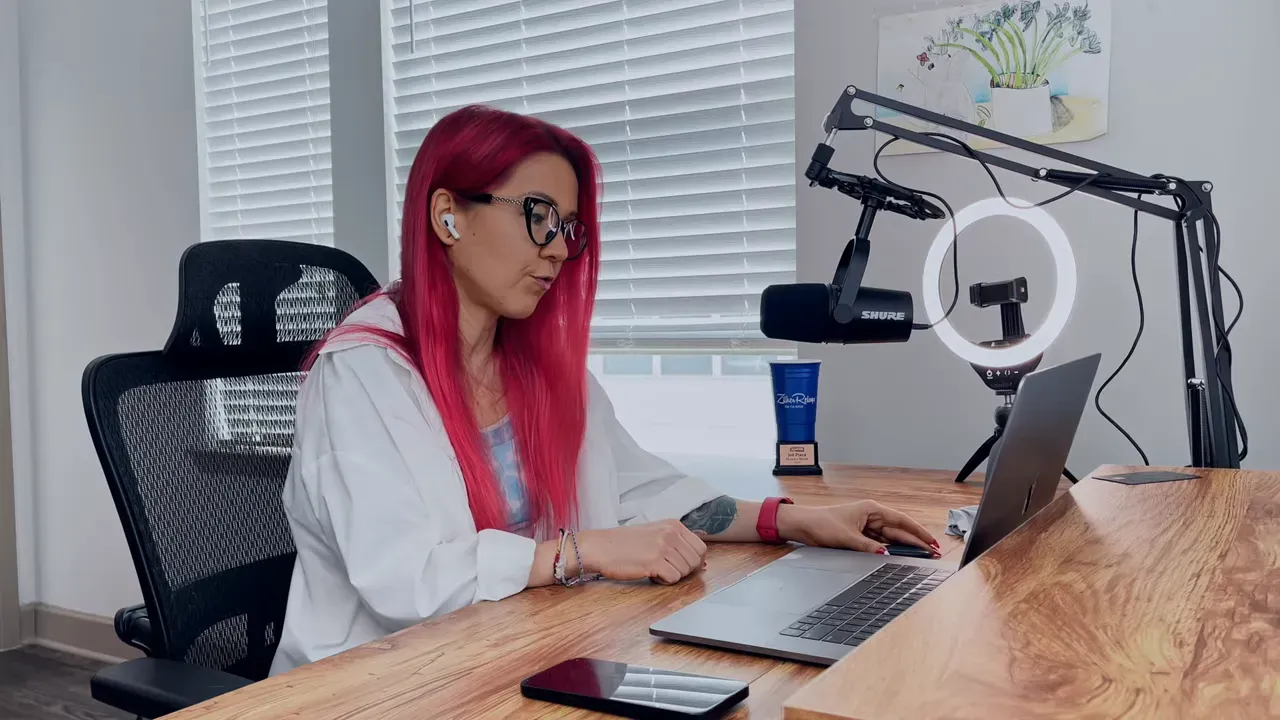 Женщина с розовыми волосами в очках за деревянным столом с ноутбуком, микрофоном и кольцевой лампой